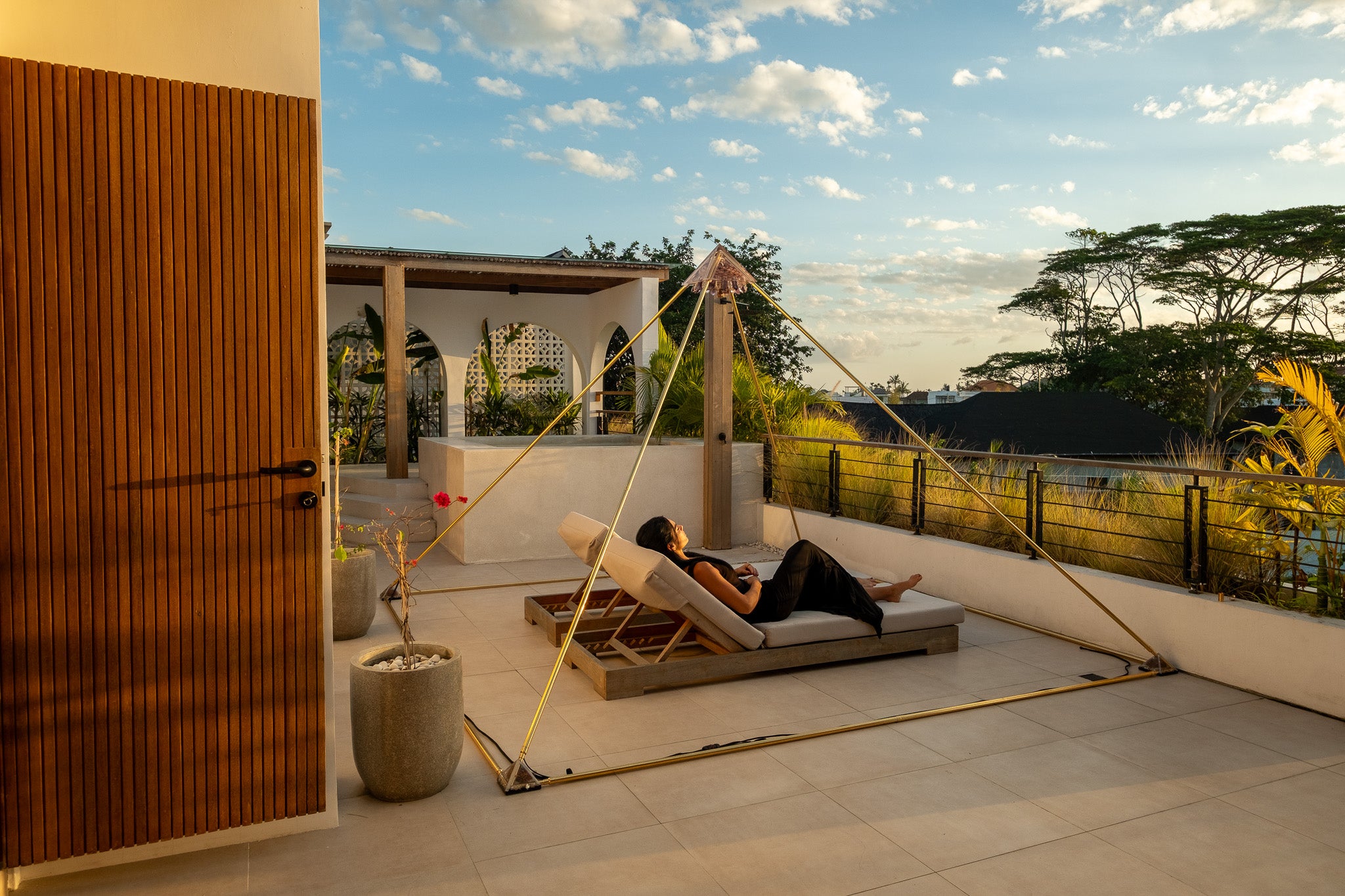 A person relaxes on a cushioned lounge chair on a modern outdoor terrace, with a large gold-framed tension fabric canopy set up nearby. The scene includes a wooden slatted door, potted plants, a patio floor, and a landscaped garden with trees under a blue sky with scattered clouds.